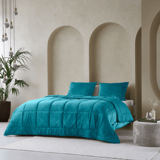 A modern bedroom featuring the Zelesta Velvetbed in petrol, with matching pillows, beige walls with three arched niches, a plant, hanging pendant lights, and a round ottoman atop a light rug.