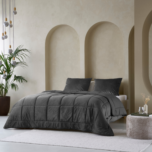 A modern bedroom showcases the Zelesta Velvetbed in anthracite, neatly made and set against a beige wall with three arched alcoves. Decorative touches include a potted plant, pendant lights, and a round ottoman.